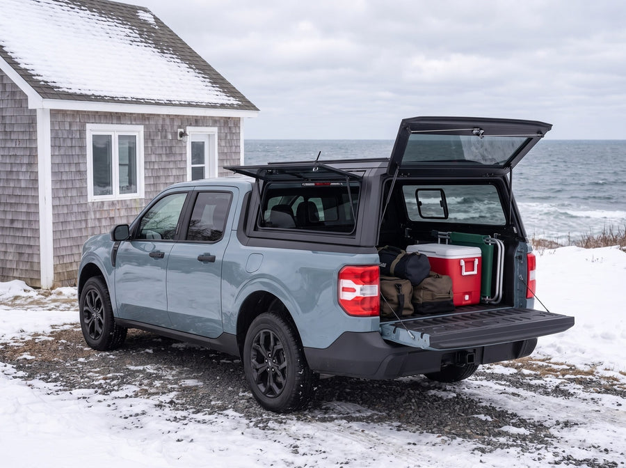 Ford Maverick truck cap open at a snow-covered winter cottage with duffel bags, a cooler and a camp chair packed in the bed