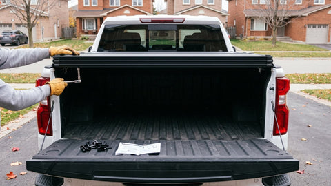 Installing a tonneau cover on a white Chevy Silverado: gloved hands tighten a clamp under the folded cover