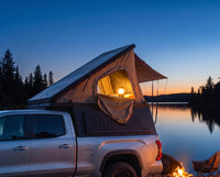 Chevrolet Colorado pop-up camper glowing at dusk beside a northern lake with a campfire