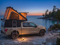 Chevy Colorado pop-up truck cap camper glowing at dusk on the Georgian Bay shore with camp chairs and a lantern
