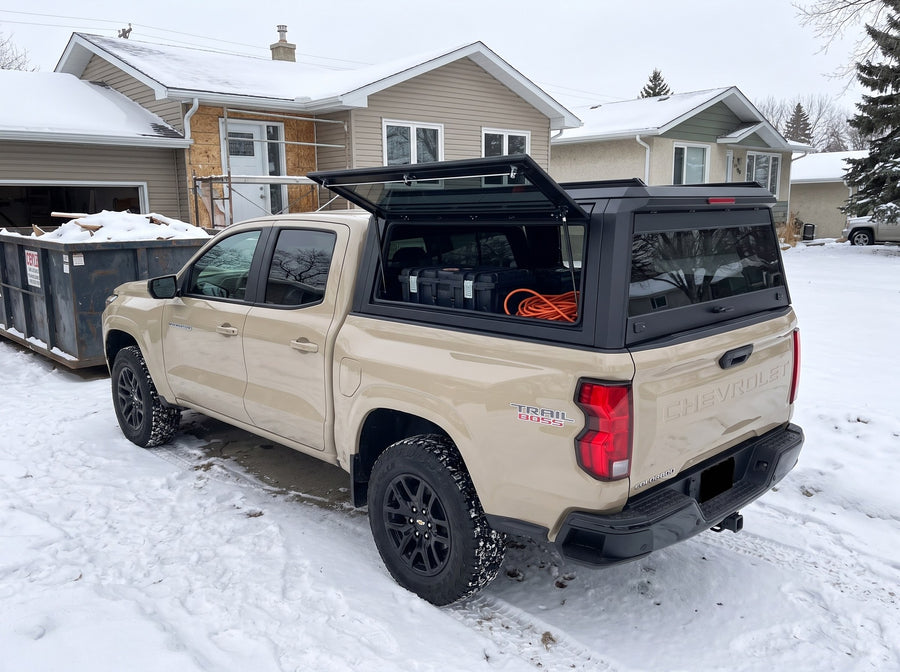 Chevy Colorado truck cap with the side window lifted open at a snowy job site in winter, tool cases and an extension cord inside