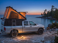 Dodge Ram 2500/3500 pop-up truck cap camper glowing at dusk on the Georgian Bay shore with camp chairs and a lantern
