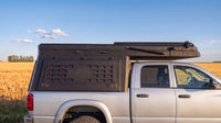 Dodge Ram 2500/3500 truck cap camper closed for driving, showing the locking side MOLLE panel and low-profile roof beside a prairie wheat field