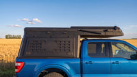 Ford F-150 truck cap camper closed for driving, showing the locking side MOLLE panel and low-profile roof beside a prairie wheat field