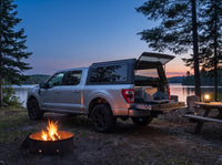 Ford F-150 with the truck cap open at a lakeside campsite at dusk, campfire and lantern lit