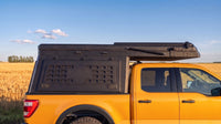 Ford Maverick truck cap camper closed for driving, showing the locking side MOLLE panel and low-profile roof beside a prairie wheat field