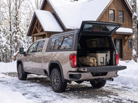 GMC Sierra 2500HD 3500HD truck cap open at a snow-covered winter cottage with duffel bags, a cooler and a camp chair packed in the bed