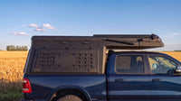 Ram 1500 truck cap camper closed for driving, showing the locking side MOLLE panel and low-profile roof beside a prairie wheat field