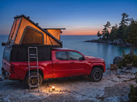 Toyota Tacoma pop-up truck cap camper glowing at dusk on the Georgian Bay shore with camp chairs and a lantern
